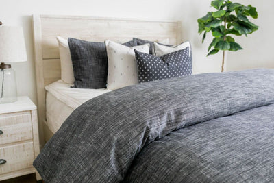 A brightly lit bedroom with a bed made with white zipper bedding and a dark gray zipper duvet folded half way down the bed and adorned with neutral black and white pillows.