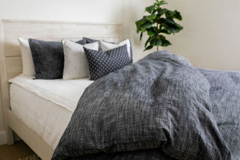 A brightly lit bedroom with a bed made with white zipper bedding and a dark gray zipper duvet partially folded half way down the bed and adorned with neutral black and white pillows.