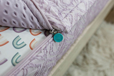 A close up view of a bed made with purple textured zipper bedding with the bed slightly unzipped to reveal the minky comforter and the rainbow pattern bottom cotton lining.