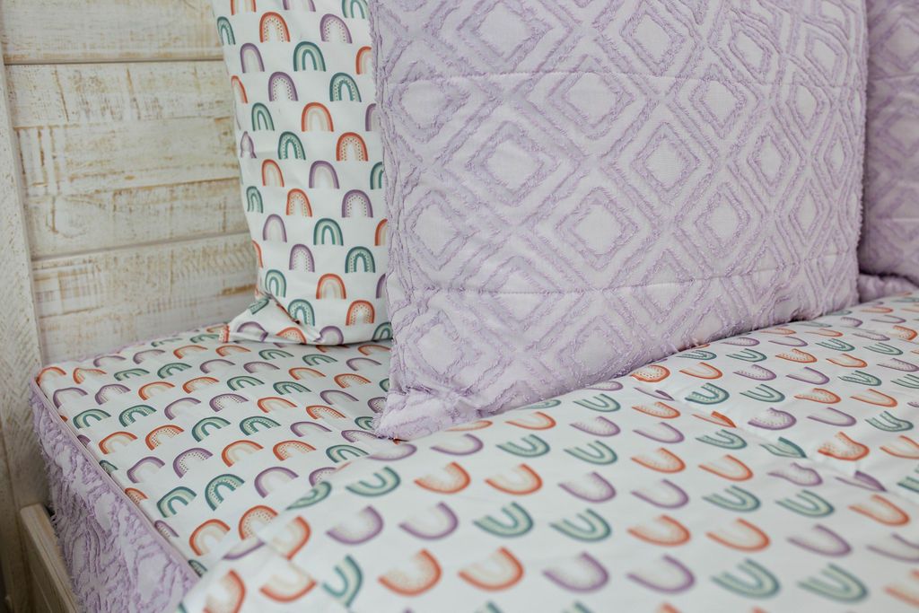A bed made with lilac zipper bedding with the comforter pulled back showing rainbows on the base sheet and pillows on top of the bed.