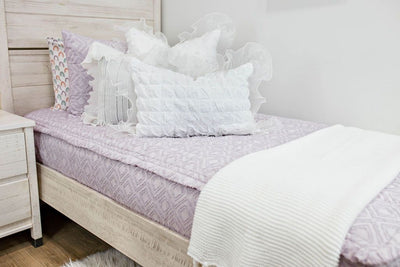 A close up view of a twin bed made with purple textured zipper bedding and white ruffled pillows and a woven blanket at the end of the bed.
