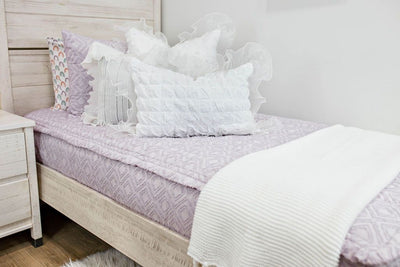 White twin bedframe with purple textured bedding, white ruffle polka dot euros, ruffle gray/blue textured pillows, white ruffle textured lumbar, and a textured white throw with braided tassels