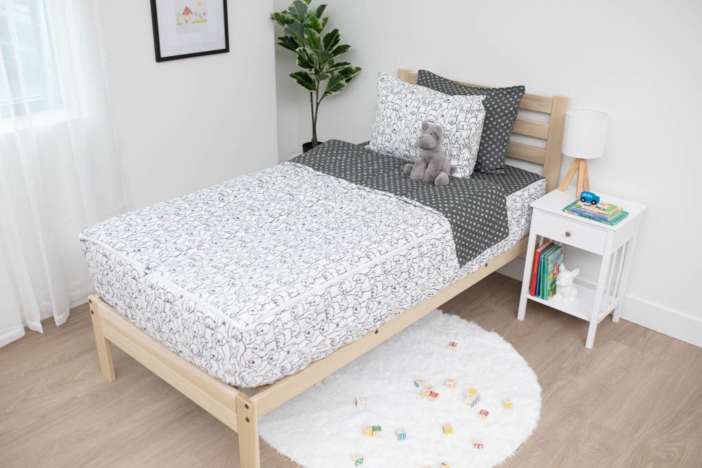 A twin bed made with charcoal and white pattern dogs kids zipper bedding, a nightstand beside it with books on the shelf and toys scattered on the floor.