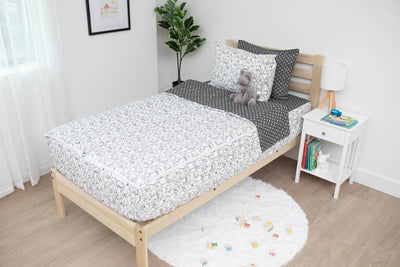 A twin bed made with charcoal and white pattern dogs kids zipper bedding, a nightstand beside it with books on the shelf and toys scattered on the floor.