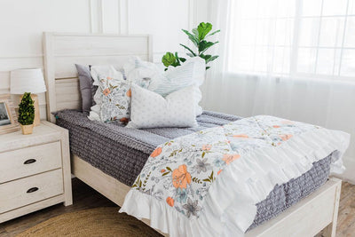 twin bed with Gray bedding with textured diamond white and blue striped euro with ruffle along the edge, floral printed pillow, white lumbar with small gray floral design and floral printed blanket with white ruffle along the edge