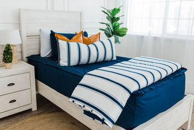 twin bed with Navy blue zipper bedding with faux leather pillows, white XL lumbar with navy vertical stripes, and white blanket with vertical navy stripes at the foot of the bed