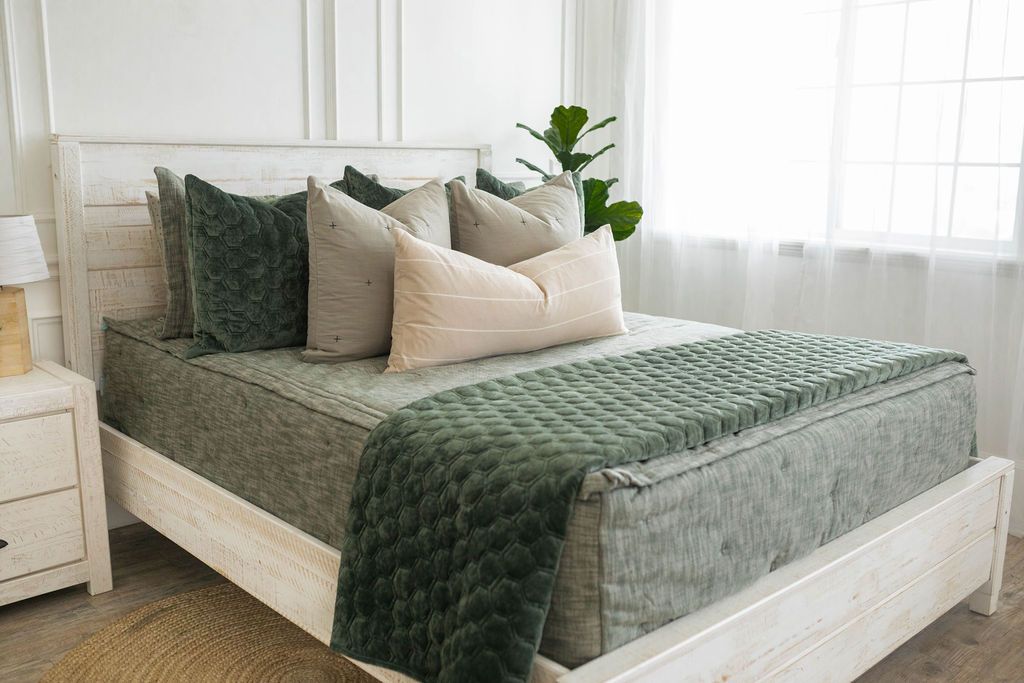 A brightly lit bedroom with a bed made with deep emerald green zipper bedding and an assortment of neutral pillows and blanket. 