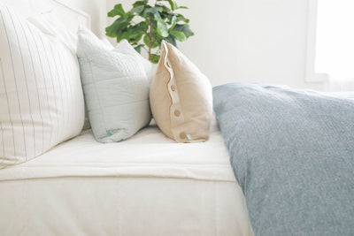 A side view of a bed made with white zipper bedding and a blue two-toned zipper duvet folded half way down the bed and adorned with neutral and light blue pillows.