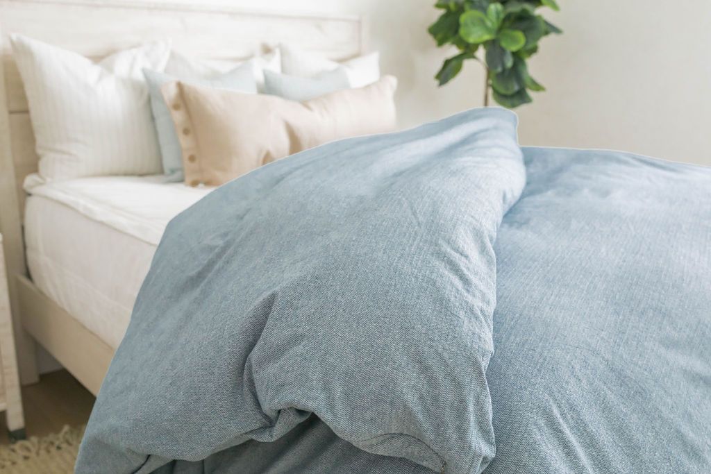 A brightly lit bedroom with a bed made with white zipper bedding and a blue two-toned zipper duvet partially folded half way down the bed and adorned with neutral pillows.