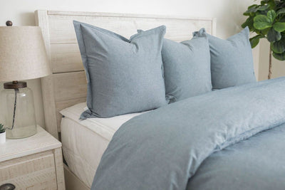 A side view of a bed made with white zipper bedding and a blue duvet with matching large blue pillows.