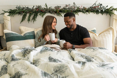 Man and woman lying underneath white and green blanket with forrest design, surrounded by cream and green pillows