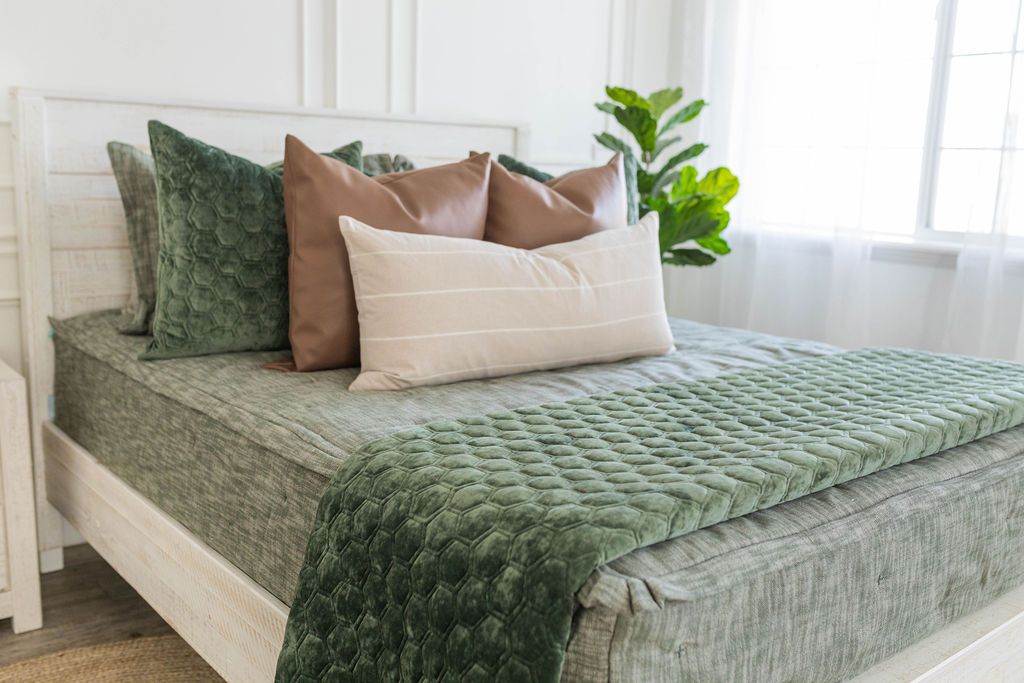 A brightly lit bedroom with a bed made with dark green zipper bedding and an assortment of neutral and leather pillows and blanket.