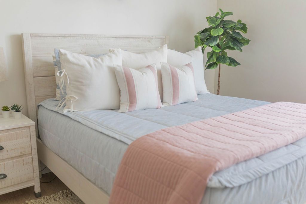 A queen bed made with light blue zipper bedding and neutral white pillows and a pink blanket at the end of the bed.