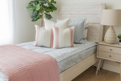 A twin bed made with light blue zipper bedding and white neutral pattern pillows and a matching pink quilted blanket.