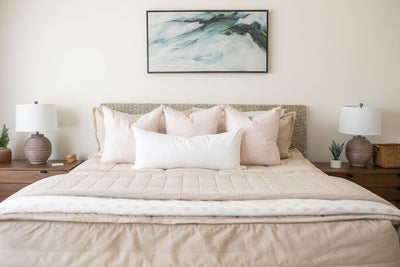 A bed made with tan zipper bedding and an assortment of neutral and light pink throw pillows and matching blankets at the end of the bed.