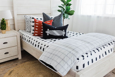 twin bed with White bedding with black dashed lines black and white striped euros, red and white dashed pillow, black lumbar with white longboard print and white and white and black grid blanket