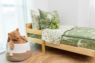 A toddler bed made with olive green dinosaur fossils kids zipper bedding and a basket of stuffed animals beside it.