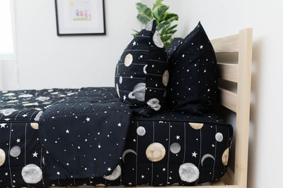A side shot of a twin bed made with black planets and stars kids zipper bedding, the top unzipped and pulled back slightly and two pillows at the head of the bed.