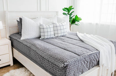 Gray bedding with textured diamond pattern and white faux fur textured euro, white and gray plaid pillow and white knitted throw with braided tassels at the foot of the bed