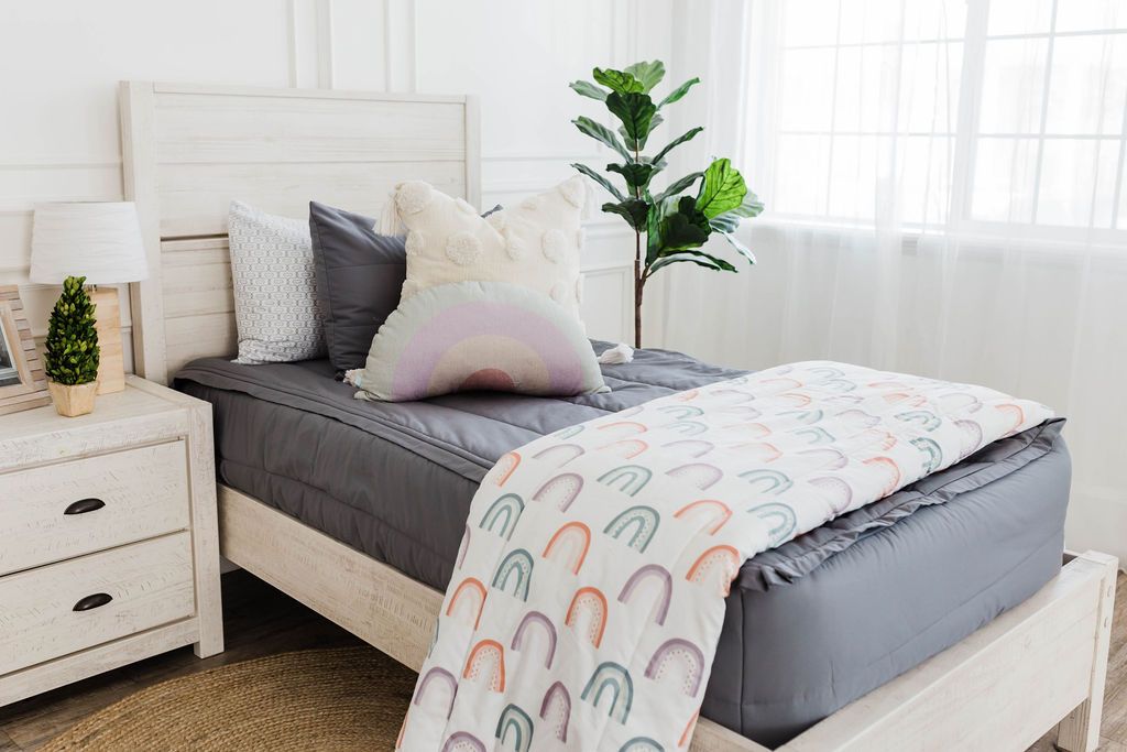twin bed with Gray zipper bedding with cream textured euro pillows with tassels, one pastel rainbow pillow, and a white blanket with ombre purple, orange and green rainbows