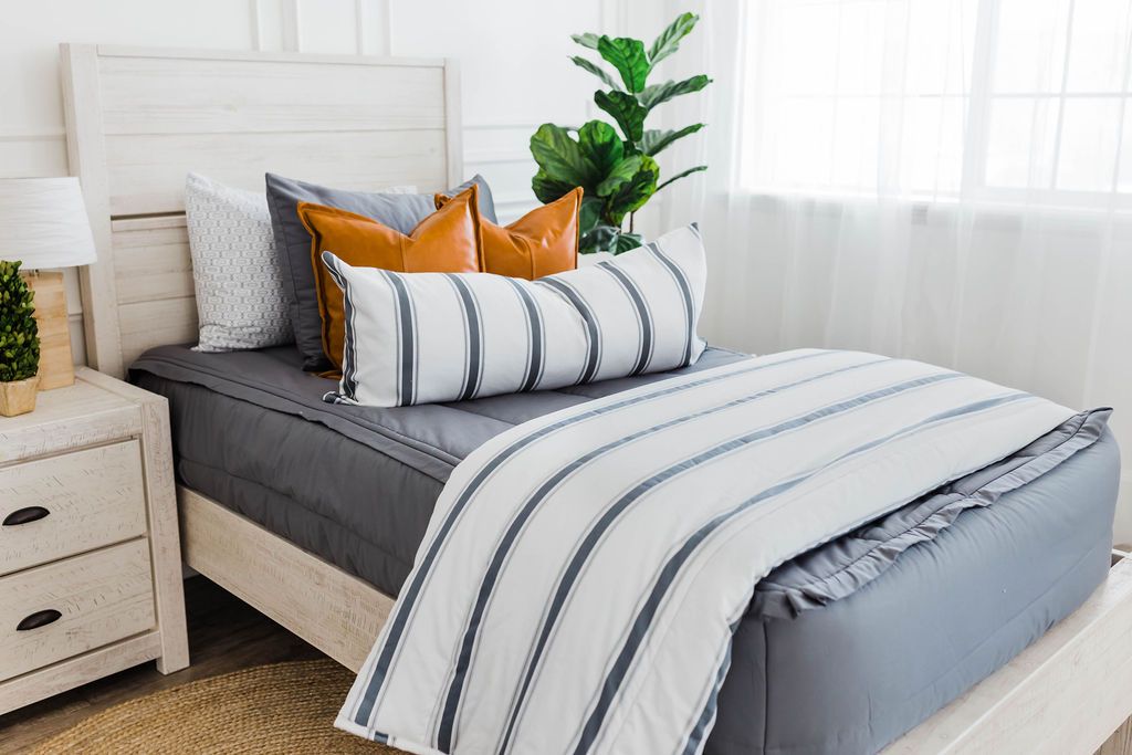 twin bed with Gray zipper bedding with faux leather pillows, white XL lumbar with gray vertical stripes, and white blanket with horizontal gray stripes at the foot of the bed