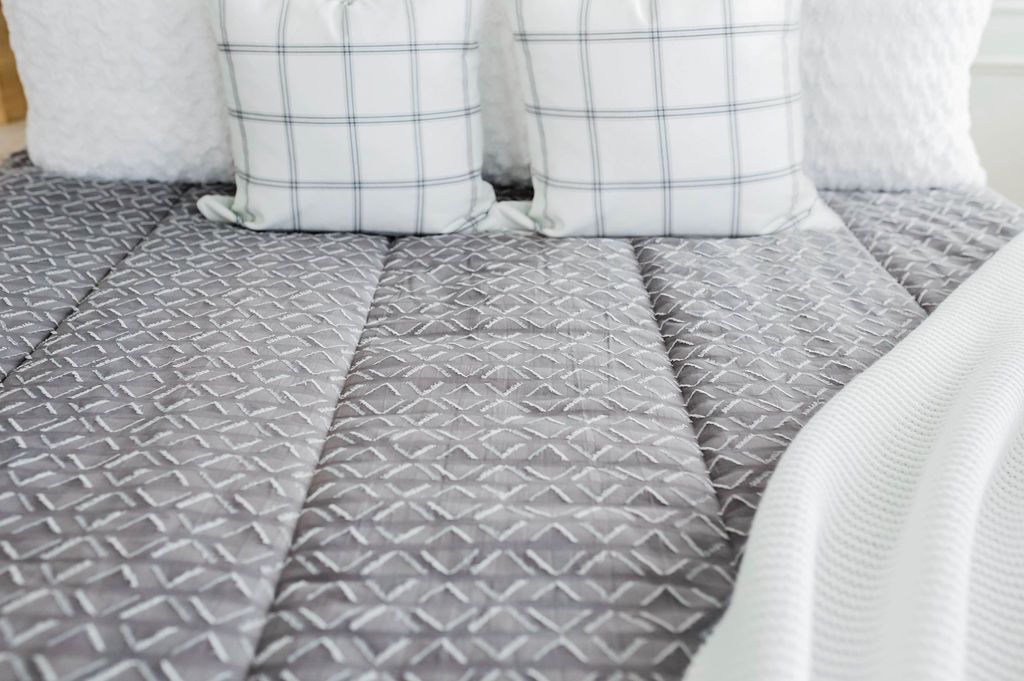 Close up of gray bedding with textured diamond pattern and white faux fur textured euro, white and gray plaid pillow and white knitted throw with braided tassels at the foot of the bed