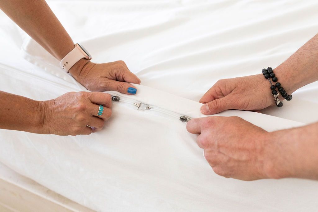 A close up view of two sets of hands zipping up a white zipper sheet set at the end of the bed. 