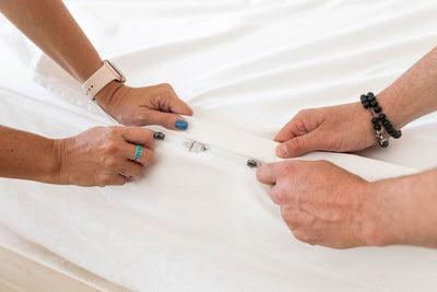 A close up view of two sets of hands zipping up a white zipper sheet set at the end of the bed. 